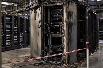 Fire-damaged server rack with disconnected cables inside a cordoned-off data center hall.